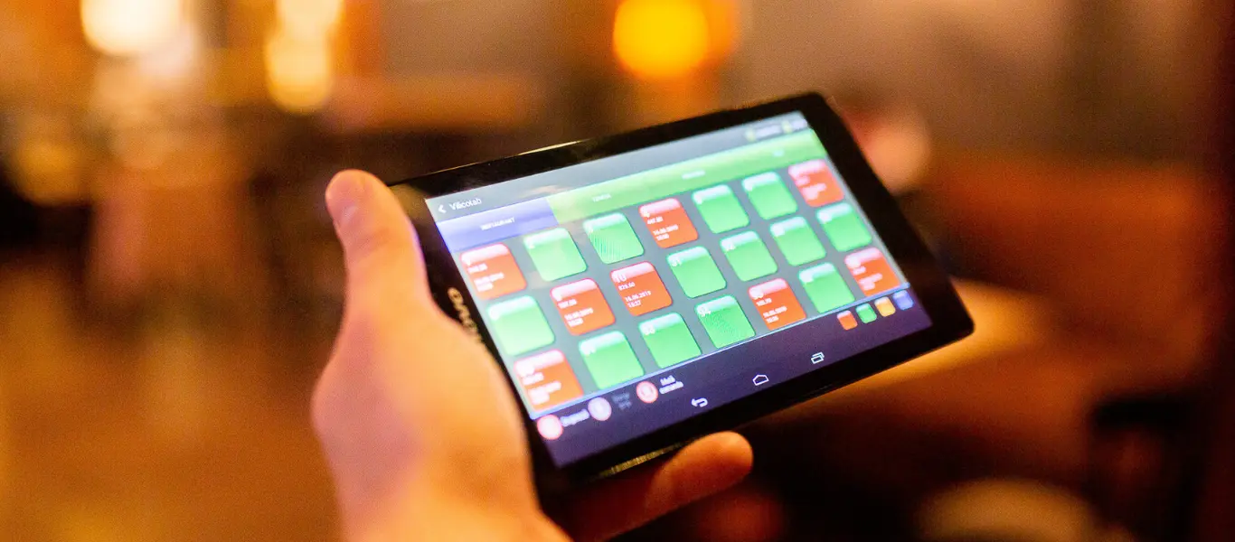 O mână ținând o tabletă cu iconițe colorate ale aplicației VilicoTab pentru ospătari pe ecran, într-un mediu slab iluminat de restaurant.